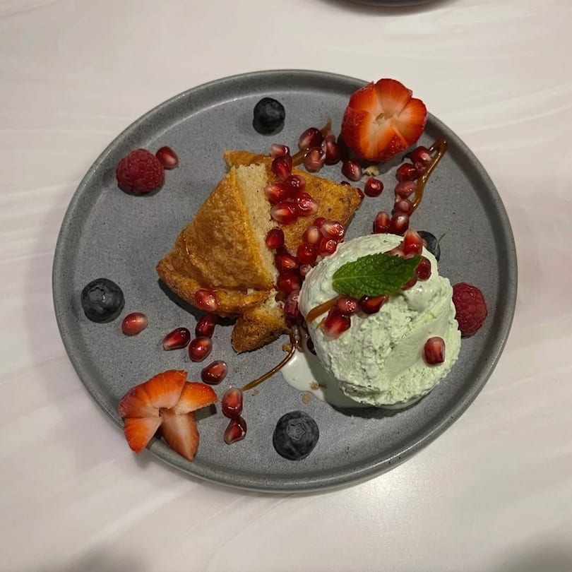 Golden cake slice with green ice cream, pomegranate seeds, and berries on a gray plate.